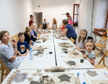 Dzieci z rodzicami siedzą przy stole i nadają kształt swoim dzwonkom wietrznym. Obok leżą wałki do ciasta.