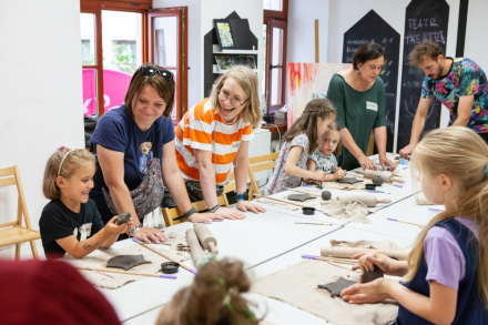Uczestnicy warsztatów stoją dookoła stołu. Lepią w glinie i ozdabiają swoje dzwonki wietrzne.