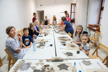 Dzieci z rodzicami siedzą przy stole i nadają kształt swoim dzwonkom wietrznym. Obok leżą wałki do ciasta.