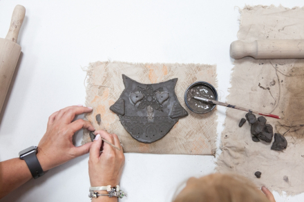 Zdjęcie z góry. Na stole leży gotowy dzwonek wietrzny w kształcie sowy. Widać dłonie mężczyzny, który rzeźbi coś w glinie.