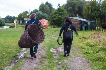 Kobieta i mężczyzna tyłem. Na plecach niosą wiklinowe gałązki oraz upleciony duży kosz.