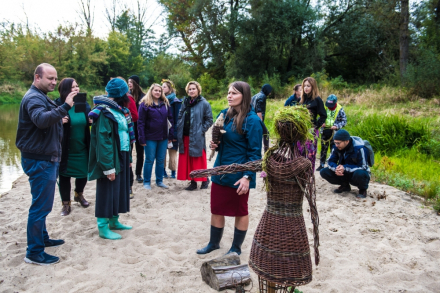 Uczestnicy warsztatów stoją dookoła kobiety, która trzyma w ręce zrobioną z wikliny rybę. Plecionka zastępuje jej mikrofon.