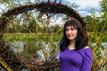 Kobieta o długich czarnych włosach stoi przy uplecionym z wikliny okręgu. Okrąg jest większy od niej.