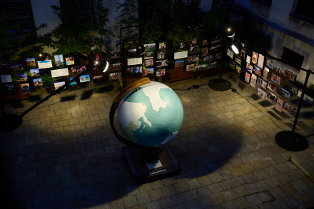 Na zdjęciu zrobionym nocą widać globus umieszczony na patio Warsztatów Kultury. Globus jest oświetlony od dołu, co sprawia, że kontynenty są wyraźnie widoczne na jasnym tle oceanów.  Z tyłu widać stojaki wystawiennicze z fotografiami i podpisami do nich. Są one oświetlone, co pozwala je oglądać i czytać nawet po zmierzchu.