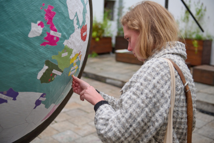 Blondynka w płaszczu w kratę przesuwa palcem po globusie, na którym zaznaczone  są miasta w różnych  krajach świata