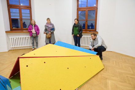 Zdjęcie przedstawia cztery osoby stojące w jasnym, przestronnym pomieszczeniu z drewnianą podłogą.  Na głównym planie znajduje się duży, trójkątny obiekt stanowiący element interaktywnej instalacji artystycznej. Obiekt jest przede wszystkim żółty, z czerwoną krawędzią i niebieskimi oraz zielonymi bokami.   Od lewej strony, pierwsza osoba to młoda dziewczyna w różowej kurtce, obok niej stoi kobieta w szarym płaszczu i różowym szaliku.  Trzecia osoba to kobieta w czarnej kurtce i zielonym swetrze,  a na końcu widzimy mężczyznę w szarym swetrze, który przykucnął obok instalacji. Wszystkie osoby są zwrócone w stronę aparatu i zdają się obserwować lub być zaangażowane w interakcję z obiektem.    W tle widoczne są duże okna.  Jest wieczór.