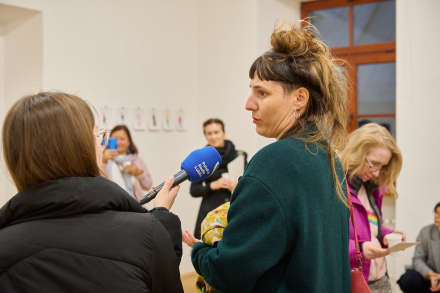 Zdjęcie przedstawia grupę osób w jasnym, przestronnym pomieszczeniu. Na pierwszym planie znajduje się kobieta w ciemnozielonym swetrze. Stoi bokiem do kamery. Udziela wywiadu. Obok niej widzimy dziennikarkę radiową, która trzyma niebieski mikrofon z logo Polskiego Radia Lublin.