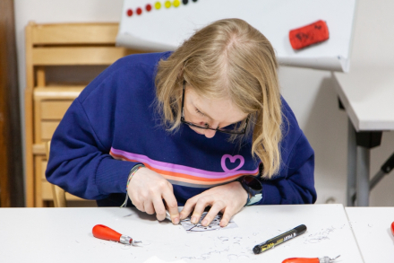 Kobieta siedzi przy stole i wycina wzór z linorytu.