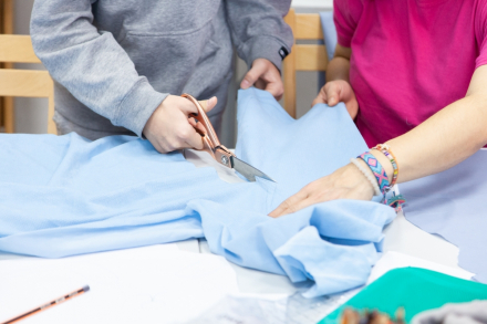 Jedna kobieta trzyma materiał, a druga kroi go nożyczkami,