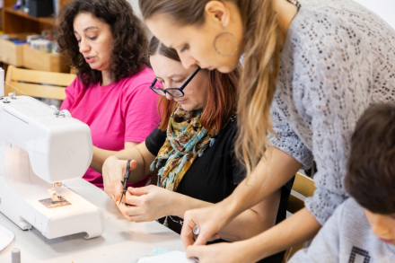 Dwie kobiety i dziewczynka szyją termofor. Na stole stoi maszyna o szycia.