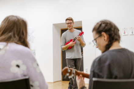 Mężczyzna chodzi po pomieszczeniu i gra na ukulele. Dookoła niego na krzesłach siedzą kursanci i uczą się grać.