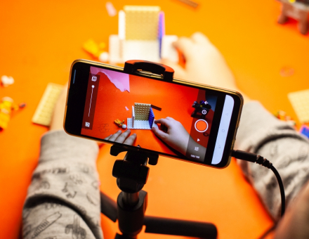 Na statywie na stole stoi telefon. Widać ręce dziecka, które buduje coś z klocków lego i robi temu zdjęcia.