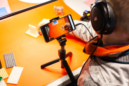 Zdjęcie z góry. Chłopiec w słuchawkach na głowie przesuwa drobne elementy po stoliku i robi temu zdjęcie.