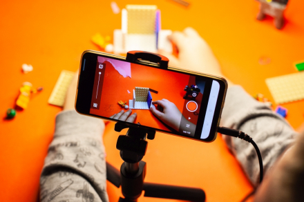 Na statywie na stole stoi telefon. Widać ręce dziecka, które buduje coś z klocków lego i robi temu zdjęcia.