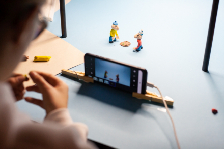 Dłonie dziecka, które lepi z plasteliny postacie Pata i Mata. Obok stoi komórka z włączonym aparatem.