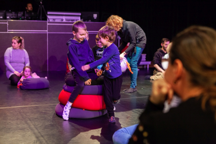 Dzieci siedzą na scenie. Pośrodku dwaj chłopcy próbują jednocześnie usiąść na stertę poduszek.