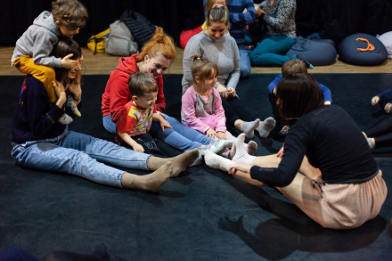 Dzieci siedzą na podłodze między nogami swoich rodziców, oparte o nich plecami.
