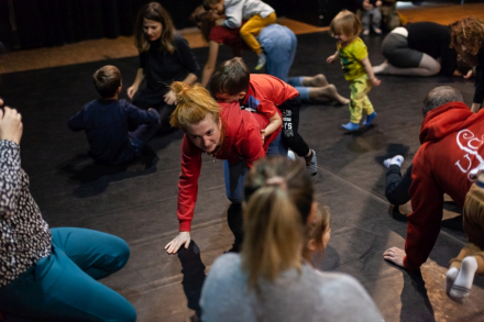 Rodzice z dziećmi siedzą do siebie twarzami. Animatorka chodzi pomiędzy nimi na czworaka.