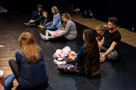 Uczestnicy warsztatów siedzą na podłodze w okręgu  i patrzą na prowadzącą.