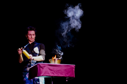 Mężczyzna trzyma w ręce zapalarkę gazową. Przed nim stoi stolik, na którym stoi podpalona miska. Z naczynia unosi się dym.