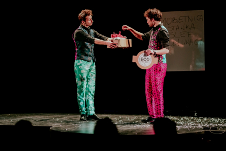 Dwaj magicy stoją na scenie. Jeden trzyma pudełko, drugi wyjmuje coś z niego. W tle znajduje się zapisana tablica.