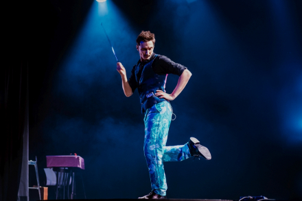 Mężczyzna stoi na jednej nodze, podpiera się pod bokiem, a drugą ręką wymachuje laską w powietrzu.