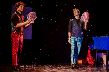 Dwaj mężczyźni stoją na scenie z kartami w rękach. Układają je w wachlarz. W tle pozostałe rekwizyty niezbędne do przedstawienia.