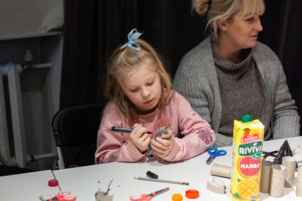 Dziewczynka rysuje coś na rolce po papierze toaletowym. Obok przy stoliku siedzi jej mama.