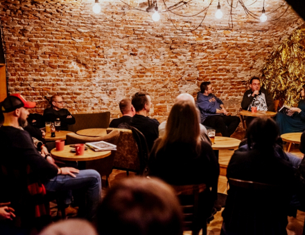 Ludzie siedzą przy stolikach i słuchają rozmowy.