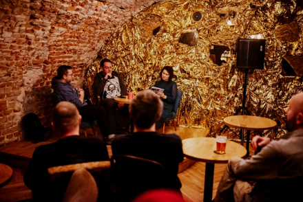 Przy stoliku siedzą Jarek Szubrycht i dwoje prowadzących. Grupa słuchaczy przygląda im się.