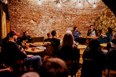 Ludzie siedzą przy stolikach i słuchają rozmowy.