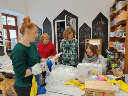 Uczestnicy warsztatu zebrani wokół stołu. Jedna kobieta przygotowuje materiał, a druga siedzi przy maszynie.