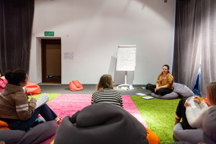 Uczestnicy warsztatu siedzą w kręgu. Prowadząca siedzi obok tablicy.