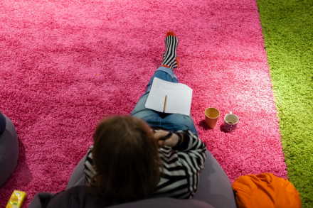 Zdjęcie zrobione z góry. Przedstawia kobietę siedzącą na pufie z zeszytem na kolanach i kubkiem herbaty obok.