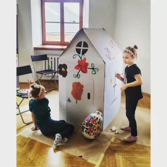 Dziewczynki ozdabiają kartonowy domek, przed którym stoi duża pisanka. 