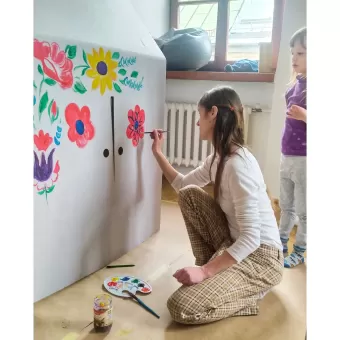 Dziewczyna maluje kwiatka na oknie kartonowego domku.