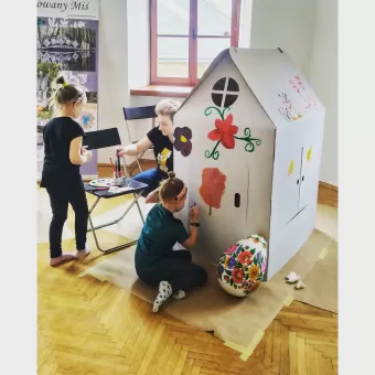 Dwoje dzieci i kobieta malują kwiaty na kartonowym domeku.