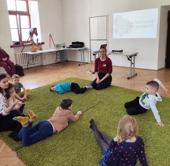 Kobieta siedzi siedzi z dziećmi na dywanie. Część dzieci siedzi, a część leży.