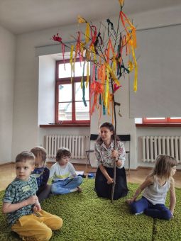 Kobieta trzyma gałąź z wstążkami, obok siedzą dzieci.