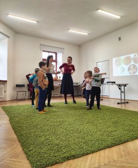 Dzieci i dwie kobiety stoją w kręgu na dywanie.
