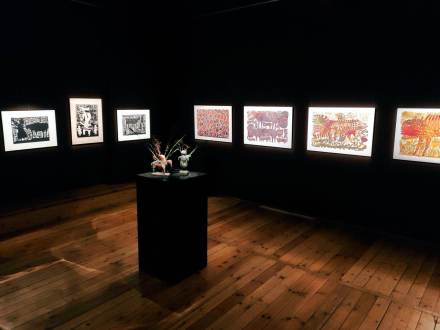 Zawieszone obrazy, oświetlone w półmroku na sali. Na środku stoi kolumna, na której są ustawione  figurki.