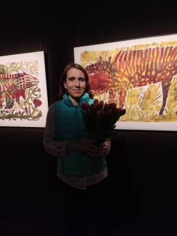 Autorka wystawy trzyma kwiaty i stoi przed swoimi dziełami.