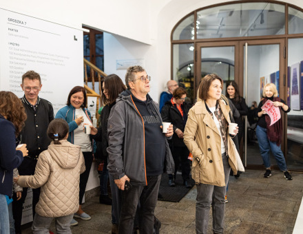 Zdjęcie przedstawia grupę osób zgromadzonych w jasnym, przestronnym korytarzu Warsztatów Kultury. Ludzie stoją w luźnych grupach, niektórzy trzymają kubki. Na pierwszym planie, po lewej stronie, widoczna jest kobieta w niebieskim płaszczu, rozmawiająca z mężczyzną i młodą dziewczyną w jasnobeżowym płaszczu.  Na środku widzimy mężczyznę w okularach i szarym płaszczu z czerwonym paskiem. Mężczyzna jest zamyślony lub wpatrzony w coś poza kadrem. Kobieta po jego prawej stronie, ubrana w beżowy płaszcz trzyma kubek i wydaje się głęboko pochłonięta wystawą.