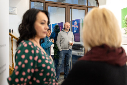 Zdjęcie przedstawia grupę osób w jasno oświetlonym korytarzu na parterze budynku Warsztatów Kultury.  Na pierwszym planie z lewej strony znajduje się kobieta w zielonej sukience w różowe kropki. Skierowana jest twarzą do aparatu. Pośrodku, w tle, stoi łysy mężczyzna w szarym swetrze, który uśmiecha się szeroko, a w ręku trzyma kubek — wygląda na zrelaksowanego i zaangażowanego w to, co się dzieje Obok niego, również w tle, znajduje się kobieta w niebieskim swetrze, trzymająca kubek, skierowana bokiem do aparatu. Z prawej strony kadru widać tył głowy kobiety z jasnymi włosami.  W szklanych drzwiach odbijają się zawieszone na ścianie kolorowe plakaty.