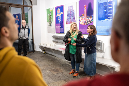 Zdjęcie ukazuje grupę osób w jasno oświetlonym korytarzu na parterze budynku Warsztatów Kultury.  Na ścianie wiszą liczne, kolorowe plansze. W centrum kadru widzimy kuratorki wystawy opowiadające o jej powstaniu i o dziełach; pierwsza z nich to blondynka, która ma na sobie czarny płaszcz, zieloną bluzkę, małą torebkę i pomarańczowe budy, druga — brunetka ubrana jest w granatową bluzę i spódnicę jeansową. W tle po prawej stronie stoi mężczyzna w okularach i niebieskiej kurtce.