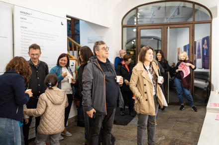 Zdjęcie przedstawia grupę osób zgromadzonych w jasnym, przestronnym korytarzu Warsztatów Kultury. Ludzie stoją w luźnych grupach, niektórzy trzymają kubki. Na pierwszym planie, po lewej stronie, widoczna jest kobieta w niebieskim płaszczu, rozmawiająca z mężczyzną i młodą dziewczyną w jasnobeżowym płaszczu.  Na środku widzimy mężczyznę w okularach i szarym płaszczu z czerwonym paskiem. Mężczyzna jest zamyślony lub wpatrzony w coś poza kadrem. Kobieta po jego prawej stronie, ubrana w beżowy płaszcz trzyma kubek i wydaje się głęboko pochłonięta wystawą.