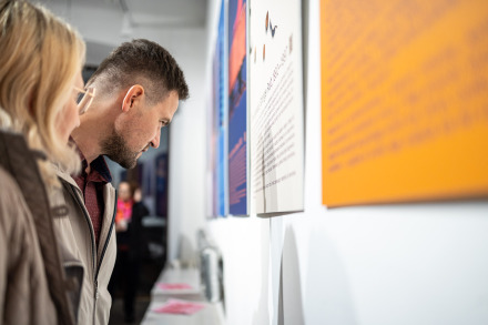 Na zdjęciu widać dwie osoby, mężczyznę i kobietę. Oglądają wystawę w korytarzu na parterze budynku Warsztatów Kultury. Zdjęcie jest wykonane z boku, skupia się na profilach obserwatorów. Mężczyzna, z lekko zarośniętą brodą i krótko przystrzyżonymi włosami, jest ubrany w jasną kurtkę i bordową koszulę we wzorek.  Kobieta, widoczna częściowo z lewej strony kadru, ma na sobie okulary i jasny płaszcz. Oboje patrzą na ścianę, na której widać plansze z tekstami i obrazami. Plansza najbliżej nich to biały panel z nadrukowanym tekstem, który jest wyraźny i czytelny. W tle  widoczna jest kolejna praca w intensywnym pomarańczowym kolorze.