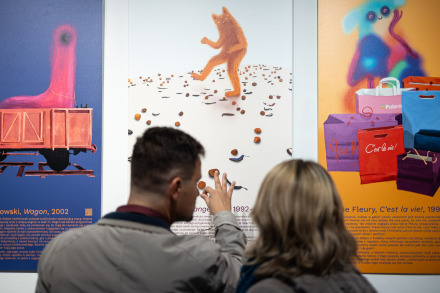 Mężczyzna i kobieta oglądają jeden z plakatów. Para wydaje się zaangażowana w rozmowę o wystawie, wskazują na pewne elementy plakatów i przyglądają im się z uwagą.  Trzy widoczne prace to: po lewej plakat z wizerunkiem czerwonej figurki na ciemnoróżowym tle, w środku plakat z pomarańczowym wilkołakiem na białym z rozrzuconymi małymi elementami wokół, a po prawej widzimy planszę z mnóstwem kolorowych toreb prezentowych i wielobarwnym misiem koalą w tle.