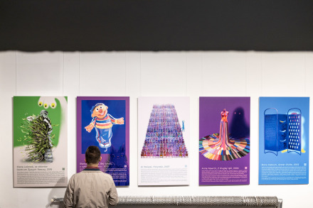 Zdjęcie pokazuje mężczyznę stojącego tyłem do kamery. Ogląda serię dużych kolorowych plansz zawieszonych na białej ścianie galerii. Każda plansza jest wyrazista i kolorowa. Na pierwszej planszy widać plastikową butelkę, z której wylewa się zieleń a nad nią widać kwiatek z żółtymi płatkami. Druga plansza to portret Erniego z Ulicy Sezamkowej na fioletowym tle. Na trzecim plakacie widać mnóstwo kolorowych krzeseł ustawionych w rzędy. Ze środkowego wyłania się tajemnicze stworzenie.  Czwarta plansza przedstawia suknię z kolorowych krawatów. Na piątym obrazie  na niebieskim tle widać dwie duże  tarki  do warzyw.