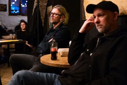 Przy stole siedzą dwaj mężczyźni. Na stoliku stoi butelka Coca-coli.